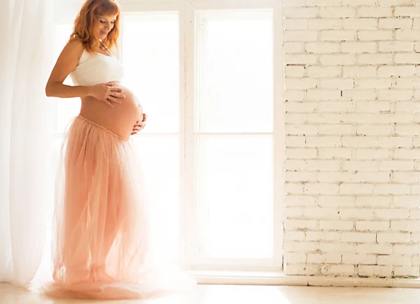 Een zwangere vrouw in een witte top en een Tule rok staat in de buurt van een groot raam — Stockfoto