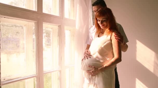 Беременная женщина и ее муж стоят у окна — стоковое видео