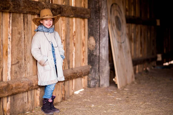 En liten flicka i en cowboyhatt står nära väggen i en lada — Stockfoto