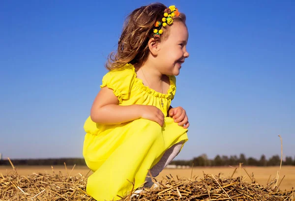 Ένα μικρό κορίτσι σε ένα κίτρινο φόρεμα κάθεται στην κορυφή του άχυρα μια — Φωτογραφία Αρχείου