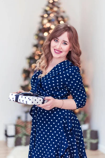 Krásná dívka v modrých šatech s dárkem v ruce. Vánoční stromek v pozadí — Stock fotografie