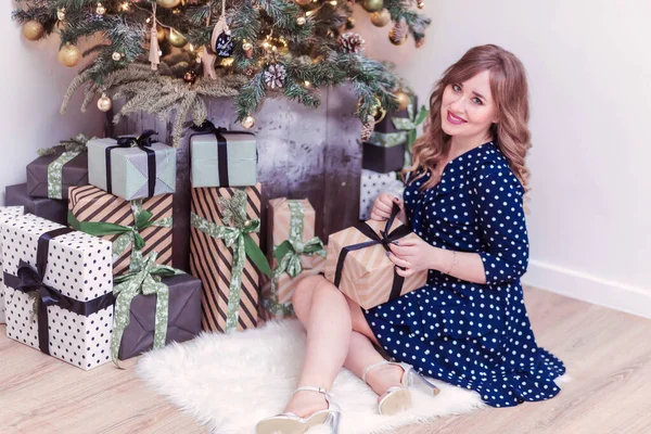 Красивая женщина в синем платье сидит рядом с новогодней елкой с подарком в руках — стоковое фото