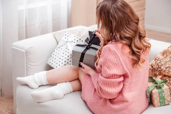 Hermosa chica en un suéter rosa y calcetines se sienta en un sofá con un regalo de Año Nuevo en sus manos —  Fotos de Stock