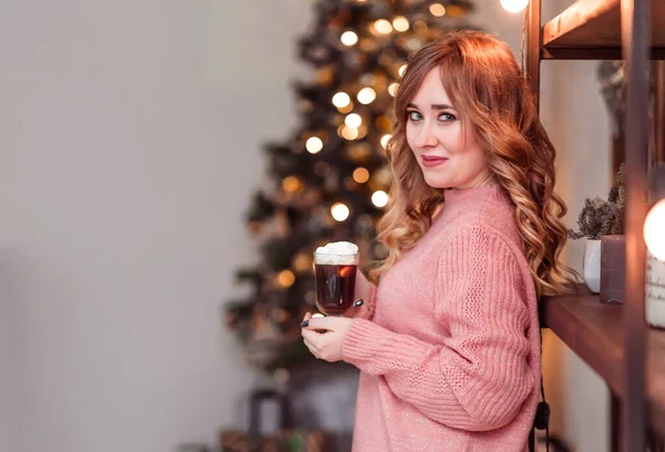 Krásná dívka v růžovém svetru s šálkem kávy a marshmallows v rukou na pozadí novoroční světla — Stock fotografie