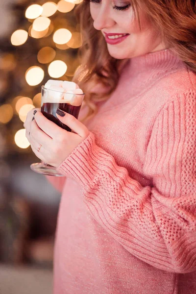 Красивая девушка в розовом свитере с чашкой кофе и зефиром в руках на фоне новогодних огней — стоковое фото