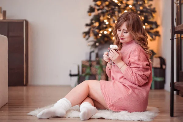 Chica sentada en una alfombra de piel cerca de un árbol de Año Nuevo con una bebida caliente en sus manos — Foto de Stock