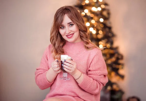 Piękna dziewczyna w różowym swetrze z filiżanką kawy i piankami w dłoniach na tle noworocznych świateł — Zdjęcie stockowe