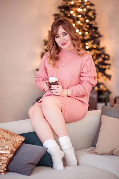 Piękna dziewczyna w różowym swetrze i skarpetkach siedzi na kanapie z filiżanką gorącego napoju w rękach — Zdjęcie stockowe