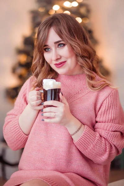 Красивая девушка в розовом свитере с чашкой кофе и зефиром в руках на фоне новогодних огней — стоковое фото