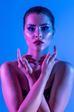 Stüdyoda renk filtreli güzel bir esmer model var. Model kollarını kavuşturdu. Neon ışıklı, mor ve mavi elbiseli güzel esmer bir kadının yakın çekimi.