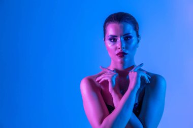 Stüdyoda renk filtreli güzel bir esmer model var. Model kollarını kavuşturdu. Neon ışıklı, mor ve mavi elbiseli güzel esmer bir kadının yakın çekimi.