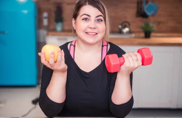 Model plus size chooses sports and healthy nutrition. Dumbbell and fruit in the hands of the model