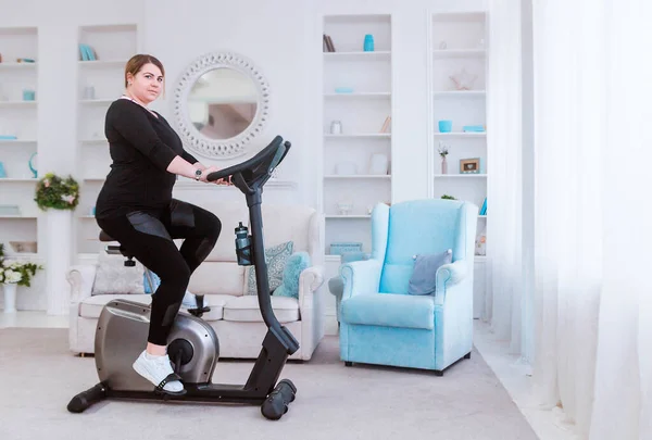 Model - a fat woman, at home engaged in a stationary bike. — Stock Photo, Image