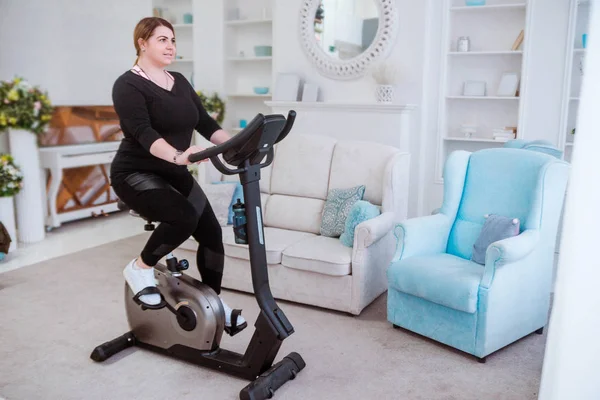 Model - a fat woman, at home engaged in a stationary bike. — Stock Photo, Image