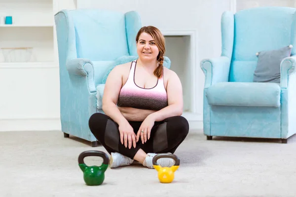 Model - dicke Frau posiert zu Hause mit Kettlebells, lächelt und blickt in die Kamera — Stockfoto