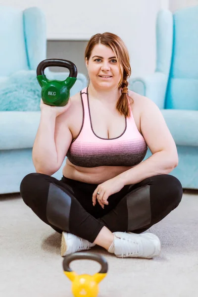 Modelo - mulher gorda posando em casa com kettlebells, sorrindo e olhando para a câmera — Fotografia de Stock