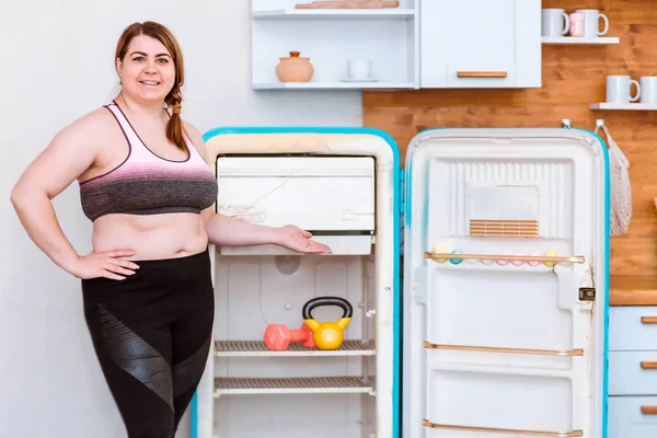 Haltere e kettlebell na prateleira do frigorífico. Mulher gorda fica ao lado da geladeira escolhendo um esporte — Fotografia de Stock