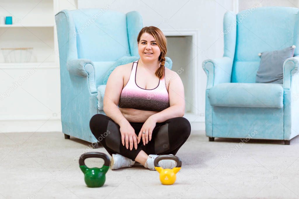 Model - fat woman posing at home with kettlebells, smiling and looking at the camera