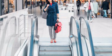 Elinde kırmızı bavul olan bir kadın bir iş merkezinde yürüyen merdivenin yanında duruyor.