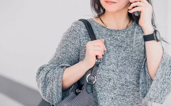 A woman in a gray dress and a large dark bag dressed over her shoulder is talking on the phone. — Stock Photo, Image