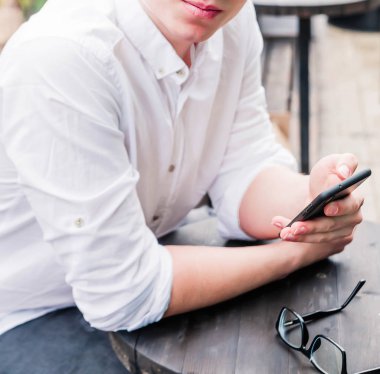 Beyaz gömlekli ve kot pantolonlu genç bir adam bir kafede bir masada oturuyor, elinde bir telefon tutuyor ve kameraya bakıyor.