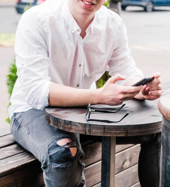 一个穿着白衬衫和牛仔裤的年轻人坐在咖啡店的桌子旁，手里拿着一部手机，看着摄像机 — 图库照片