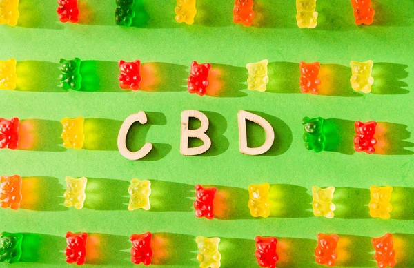 Letras de madera CBD sobre fondo verde y gelatina en forma de osos. Vista desde arriba. La sombra del sol cae sobre el fondo — Foto de Stock