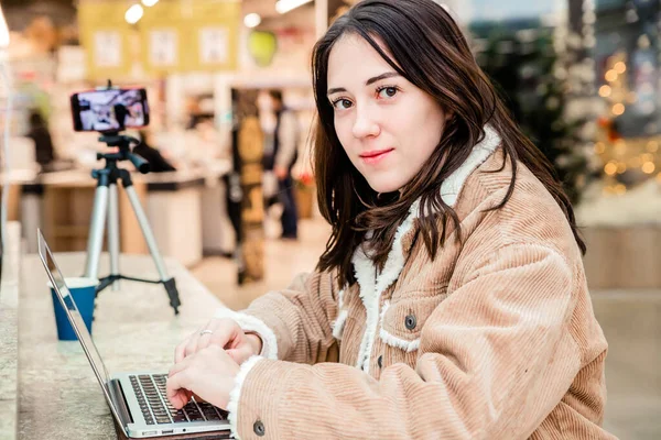Junge Bloggerin in der Mall arbeitet an einem Laptop, ein Stativ mit Mobiltelefon ist in der Nähe — Stockfoto