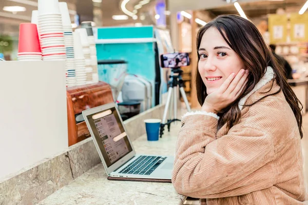 Молодая женщина блоггер в торговом центре работает на ноутбуке, штатив с мобильным телефоном рядом — стоковое фото
