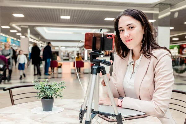 A young woman blogger in a shopping center works at a laptop, conducts live broadcasts, records videos, communicates with followers of her social networks — Stock Photo, Image