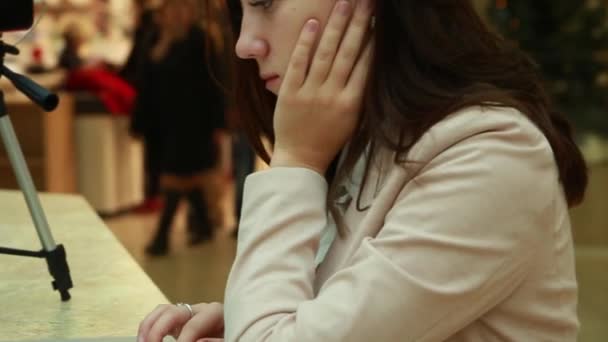 Blogger joven en el centro comercial trabajando en un portátil, un trípode con un teléfono móvil están cerca — Vídeo de stock