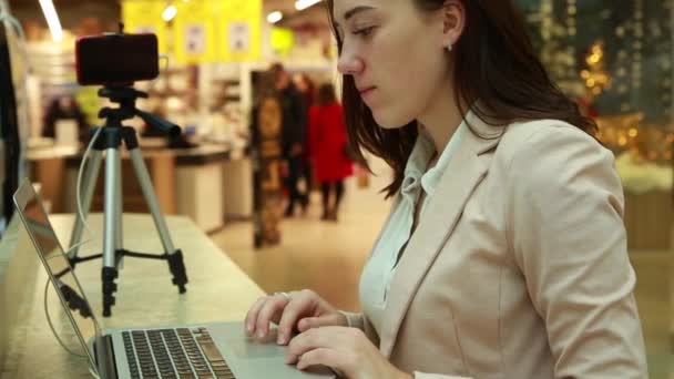 Blogger joven en el centro comercial trabajando en un portátil, un trípode con un teléfono móvil están cerca — Vídeo de stock