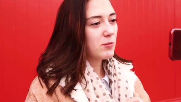 한 젊은 여성 블로거가 빨간 벽 앞에 서서 휴대폰 화면을 보고 비디오를 찍습니다. — 비디오