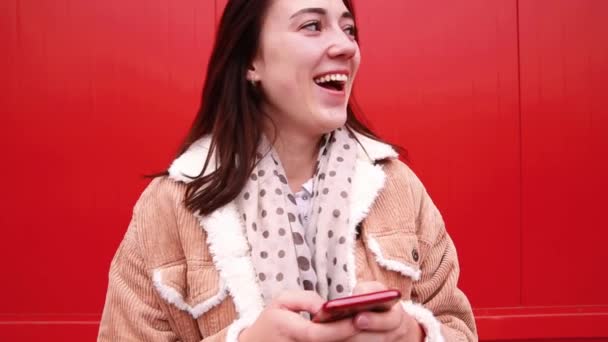 Молода жінка-блогер стоїть перед червоною стіною, дивиться на екран мобільного телефону і радіє — стокове відео