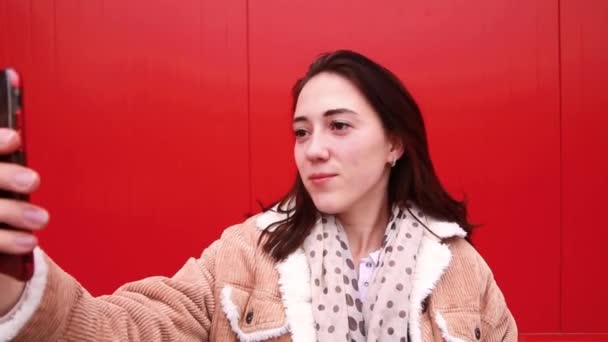 Una joven bloguera se para frente a la pared roja, mira la pantalla de un teléfono móvil y graba video — Vídeo de stock