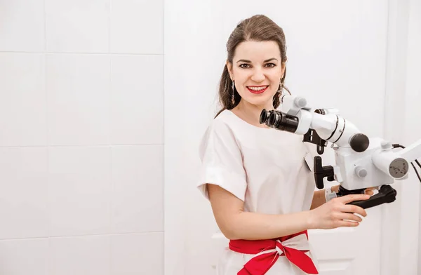 La atractiva dentista joven en traje blanco usa un microscopio en el trabajo. Doctor mirando a la cámara y sonriendo . — Foto de Stock