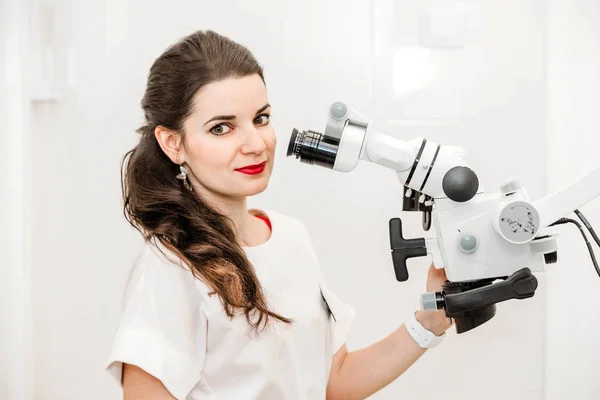 Atractiva dentista joven en un traje blanco utiliza un microscopio en el trabajo — Foto de Stock