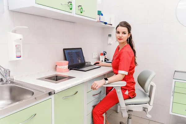 Una joven dentista atractiva se sienta en una mesa y trabaja en una computadora portátil donde los resultados del examen son visibles . —  Fotos de Stock