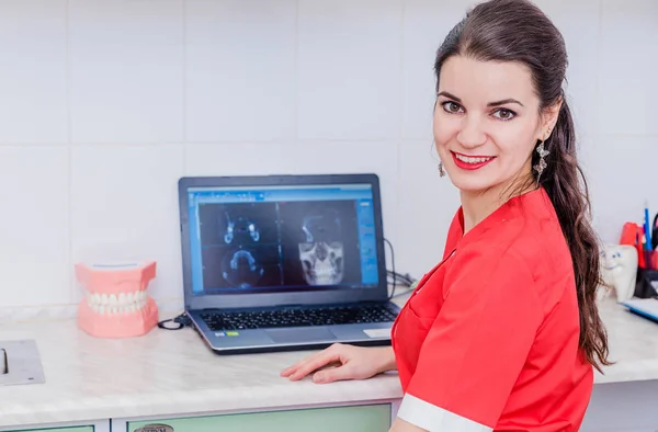 Una joven dentista atractiva se sienta en una mesa y trabaja en una computadora portátil donde los resultados del examen son visibles . — Foto de Stock