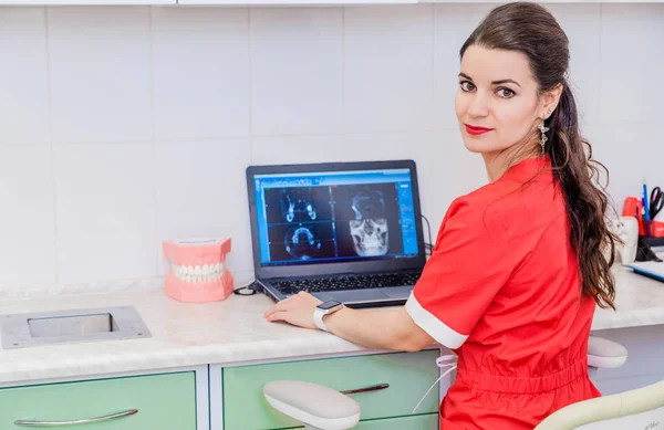 Una joven dentista atractiva se sienta en una mesa y trabaja en una computadora portátil donde los resultados del examen son visibles . —  Fotos de Stock