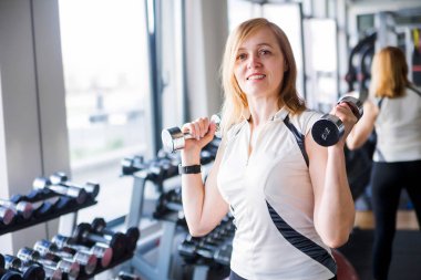 Elinde dambıllar olan yaşlı bir kadın pencerenin yanındaki spor salonunda poz veriyor.