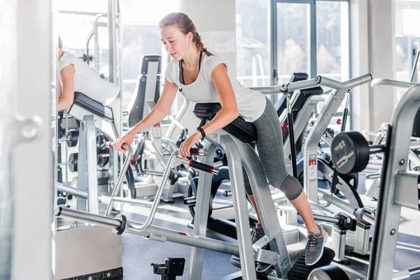 Joven hermosa chica buena forma vestida con ropa deportiva está haciendo ejercicio en el equipo deportivo en el moderno gimnasio lleno de luz solar —  Fotos de Stock