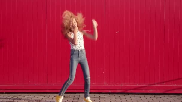 Curly blonde girl in a polka-dot print T-shirt and blue jeans dancing, fooling around and pretending to dive against a red wall. Video — Stock Footage Curly Blonde Girl Polka Dot Print Shirt Blue Jeans Dancing Royalty Free Stock Footage