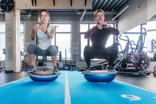 Ajuste pareja trabajando en la bola de bosu en el gimnasio. Haciendo un ejercicio en cuclillas — Foto de Stock