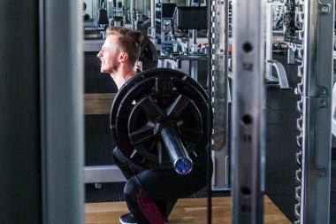 Beyaz bir adam kapalı spor salonunda halterle çömelme gösterisi yapıyor..