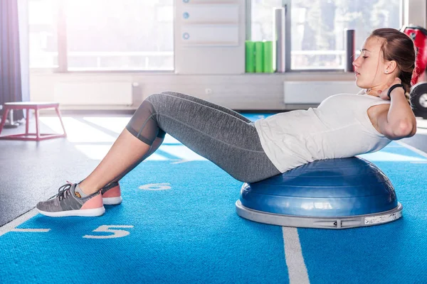 Focado esportivo bela jovem atlética morena em leggings cinza e t-shirt branca trabalhando no ginásio fazendo exercícios para músculos abdominais no treinador de equilíbrio bosu, deitar-se na bola de fitness, indoor — Fotografia de Stock