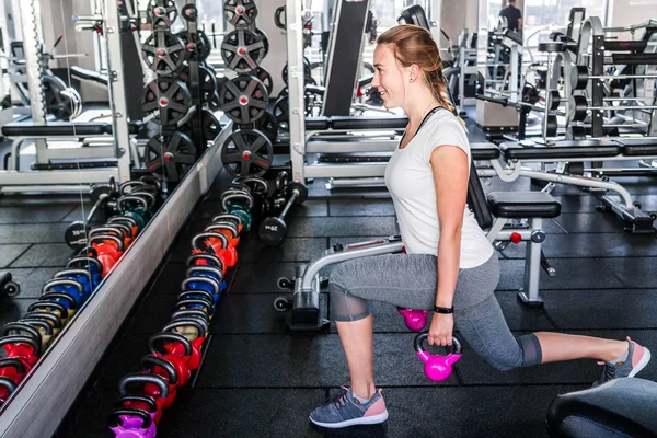 Chica en una camiseta blanca y polainas grises hace ejercicios con pesas en el gimnasio . — Foto de Stock