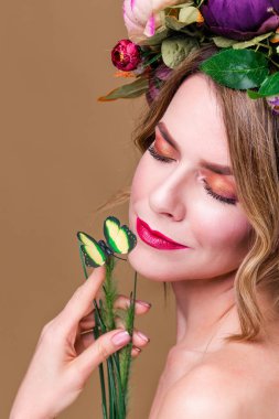 Yapay yeşil kelebekli çiçek çelengi içinde poz veren genç bir kadın, yeşil arka planda izole edilmiş.