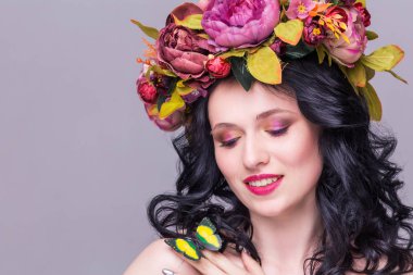 Yapay yeşil kelebekli çiçek çelengi içinde poz veren narin, genç esmer kadın, menekşe arka planda izole edilmiş.