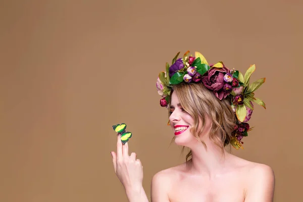 Нежная молодая женщина позирует в цветочном венке с искусственной зеленой бабочкой, изолированные на охетном фоне — стоковое фото